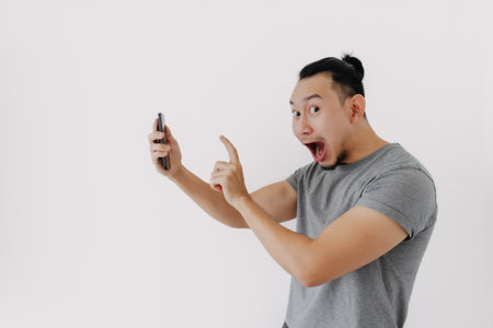 Shock and wow face Asian man in grey t-shirt use smartphone app on white.の写真素材