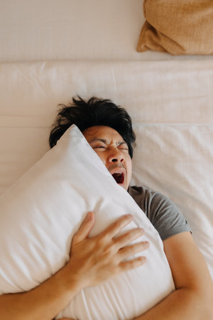 Sleepy asian man keep on sleeping on the bed, using pillow covering his face.の写真素材