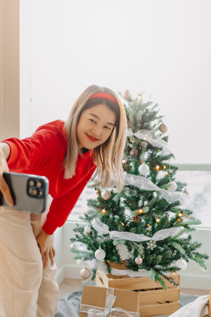 Happy and cheerful Asian woman in red sweater shirt taking selfie photo with her decorated Christmas tree in the apartment in Christmas holiday.の写真素材