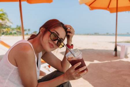 Happy Asian woman enjoy the iced tea under orange umbrella and blue sky beach.の写真素材