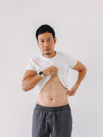 Muscular Asian man lifting shirt to display toned abdominal muscles against white wallの写真素材