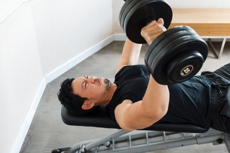 Focused male lifting heavy weights while lying on bench indoorsの写真素材