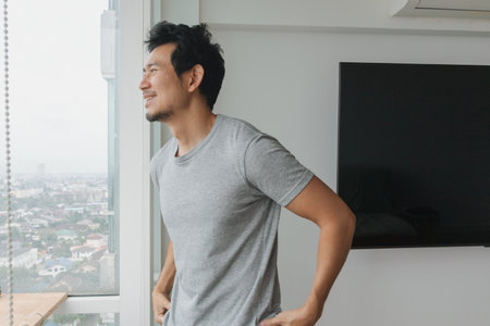 Asian male in grey t-shirt looking out with hands on waist in modern homeの写真素材