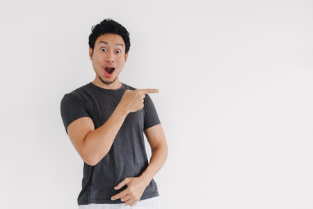 Thai male in casual outfit showing surprised pointing gesture against white backgroundの写真素材