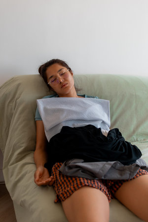 Young tired Asian female in apron leans back on sofa with pile of clothesの写真素材