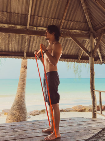 Man exercise and workout with rubber band in the hut by the sea. Beach relaxed vibe in Asia.の写真素材