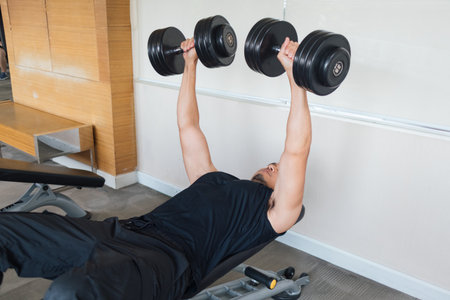 Man lifting two dumbbells while lying on flat bench inside gymの写真素材