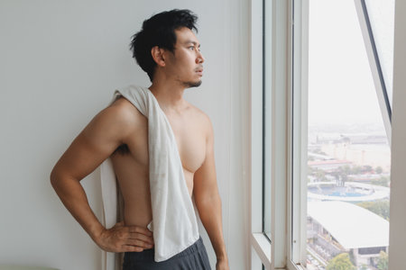 Shirtless Asian male with towel leaning on glass looking over city from apartmentの写真素材