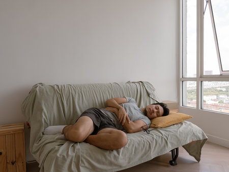 Asian male adult in casual clothes sleeps in a fetal position on the sofa, resting face on hands in...