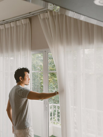 Young Thai man of Asian ethnicity opening sheer white curtain while standing by a large window...