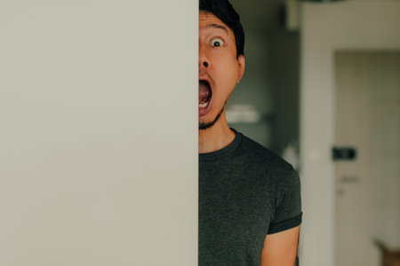 Young Thai male peeks around white door edge with open mouth and big eyes, expressing exaggerated surprise and humor in a candid pose inside a brightly lit modern apartment with casual atmosphere.の写真素材
