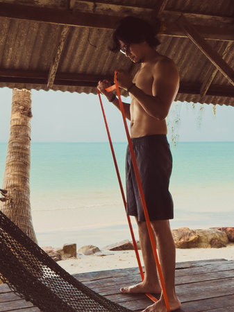 Man exercise and workout with rubber band in the hut by the sea. Beach relaxed vibe in Asia.