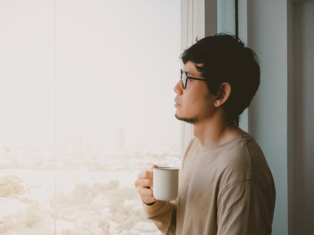 Man in glasses is holding a white coffee cup and looking out the window. The scene suggests a moment...