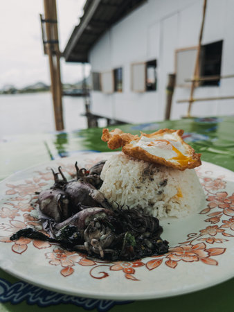 Close-up of authentic Thai cuisine featuring white rice and seasoned fried insects, served with a crispy egg on a decorative floral plate, captured on an outdoor table with a house and river background in natural light.の写真素材