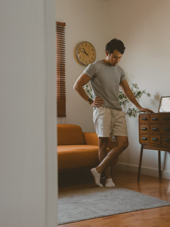 Young Thai man stands casually with both hands on his waist in a warm-toned apartment dining area, featuring soft light and wooden furnishings in a quiet domestic setting.の写真素材