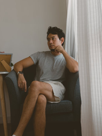 Thai male in casual outfit sitting with thoughtful expression near curtain in soft natural light and...