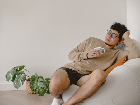 A young man is lounging comfortably at home, savoring a peaceful moment while sipping a warm cup of...