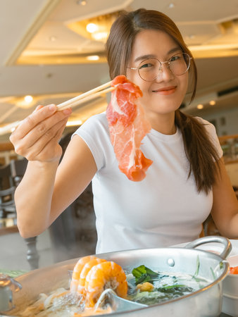 Young Thai woman with glasses smiling while holding raw beef for shabu hot pot in restaurant setting, Asian ethnicityの写真素材