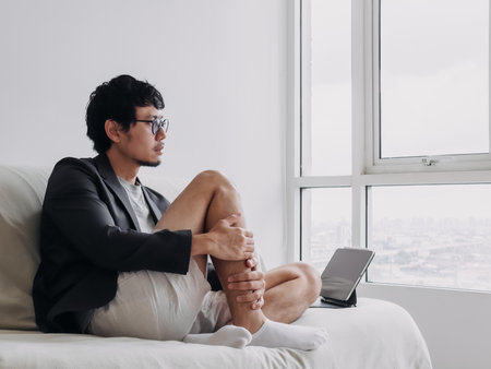 Man in a suit and glasses is sitting on a couch with his legs crossed and a tablet in front of him. He is in a relaxed and comfortable moodの写真素材