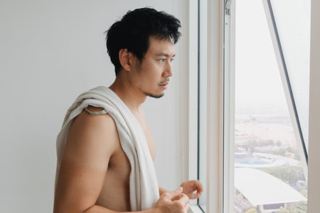 Asian shirtless male standing with towel on shoulder looking outsideの写真素材