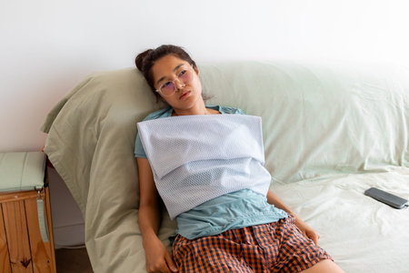 Tired Asian female wearing glasses and apron rests on sofa after houseworkの写真素材