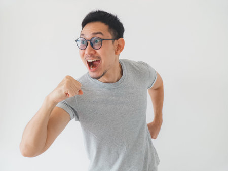 Cheerful asian man pumping fist and smiling with bright energy, casual outfit and glasses in studio...
