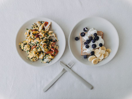 Two plates breakfast with toast and scrambled eggs, crossed fork detail on linen, vibrant tomato and...