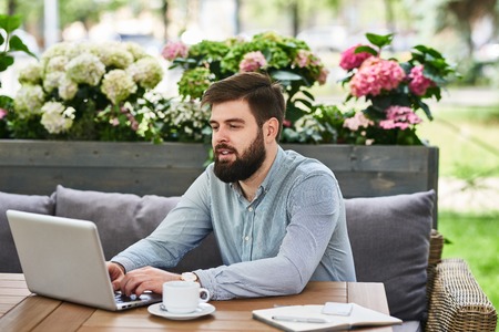 Portrait of contemporary bearded man using laptop at table in comfortable cafe lounge outdoorsの写真素材