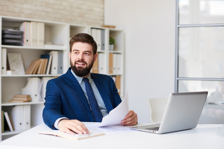 Portrait of successful  bearded businessman working at desk in light office looking away pensively and smilingの写真素材