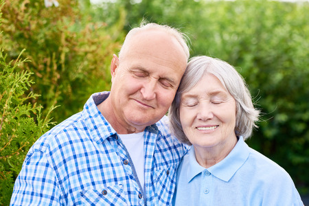 Portrait of senior couple in love, embracing leaning on each other with eyes closed and smiling standing in beautiful green garden against treesの写真素材