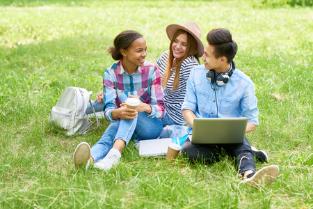 Portrait of happy students sitting on green lawn with laptop chatting and smiling enjoying break on summer dayの写真素材