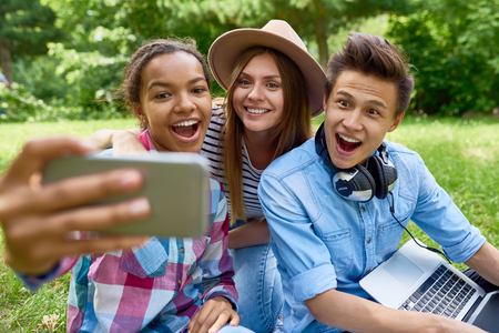 Multi-ethnic group of students taking selfie on green lawn outdoors, smiling happily looking at smartphone cameraの写真素材