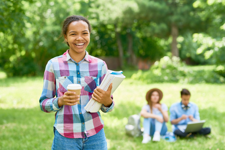 Portrait of happy mixed race student standing outdoors on green lawn  holding books and smiling looking at camera with other people in backgroundの写真素材