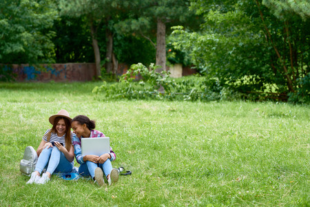 Portrait of two girls sitting on green lawn with laptop, using smartphone enjoying sunny summer dayの写真素材