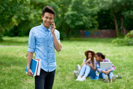 Portrait of happy Asian  student speaking by smartphone standing outdoors on green lawn  holding books and smiling looking at camera with other people in backgroundの写真素材