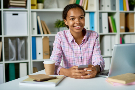 Portrait of smiling African girl studying in school library, looking at camera and smiling holding smartphoneの写真素材