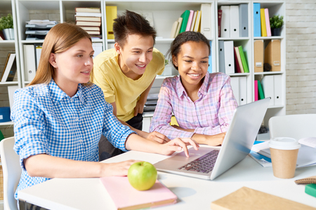 Portrait of three students using laptop studying together in library looking at screen and smilingの写真素材
