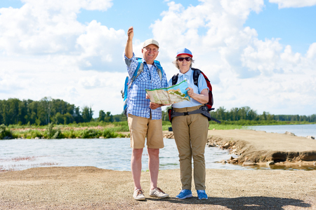 Portrait of active senior couple travelling on hiking trip, man and woman both wearing backpacks smiling at camera holding map against beautiful nature sceneryの写真素材