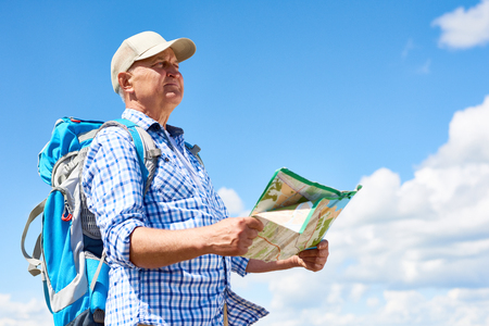 Side view portrait of active senior man travelling on hiking trip wearing backpack, looking away holding map against clear blue skyの写真素材