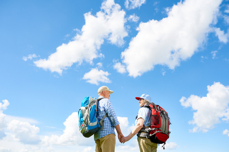 Back view portrait of active senior couple travelling on hiking trip, both wearing backpacks, standing holding hands against clear blue skyの写真素材