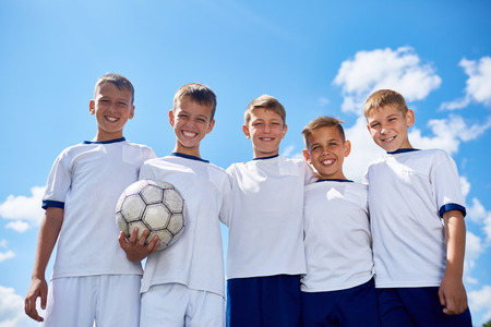 Portrait of junior football team posing smiling at camera with ball against clear blue sky in sunlightの写真素材