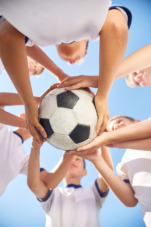 Low angle view of boys in junior football team holding ball together against clear blue skyの写真素材