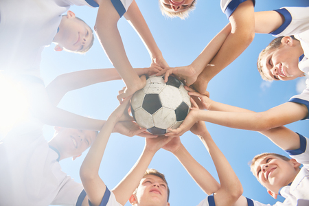 Low angle view of boys in junior football team standing in circle holding ball together against clear blue sky