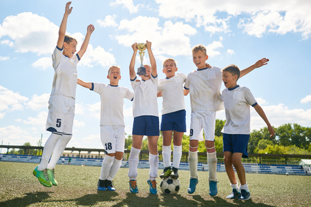 Portrait of junior football team cheering happily and jumping holding trophy cup after winning matchの写真素材