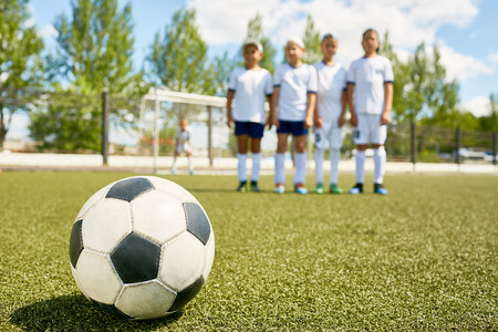 Closeup of football ball lying on green grass with junior team in backgroundの写真素材