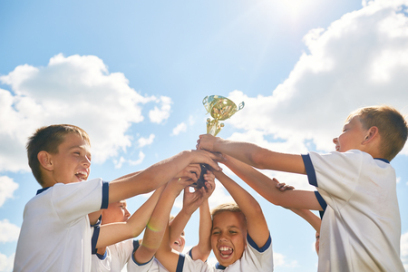 Team of children celebrating victory in sports, cheering and holding golden cup against clear blue skyの写真素材