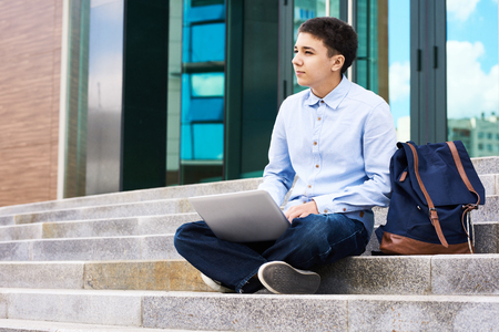 Portrait of thoughtful teenage boy sitting on school stairs outdoors using laptopの写真素材