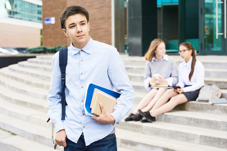 Portrait of teenage boy holding books standing outside school building, looking away pensivelyの写真素材