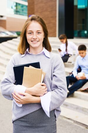 Portrait of happy teenage girl with red hair and freckles standing outside school building holding books and looking at cameraの写真素材