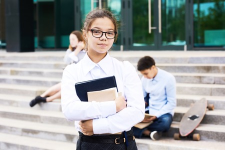 Portrait of smart teenage girl holding books standing outside school building and looking at cameraの写真素材